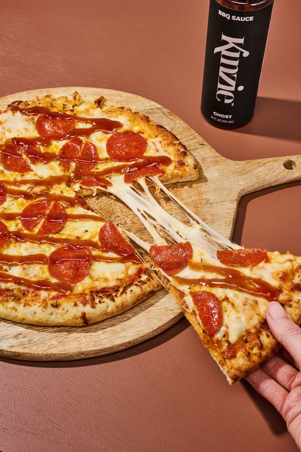 Pepperoni pizza on a wooden board with a hand picking up a slice, and a bottle of Krieger BBQ sauce in the background.
