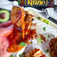 Hand holding a crispy fried item with red sauce over a burrito, with Kinzie hot sauce in the background.