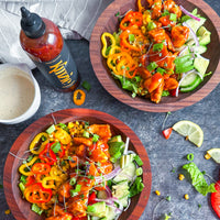 Two wooden bowls of salad with colorful vegetables and a bottle of hot sauce on a dark surface.