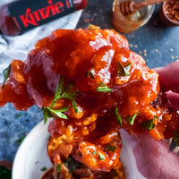 Hand holding crispy fried chicken with red sauce and green herbs, with a bottle of Ketchup in the background.