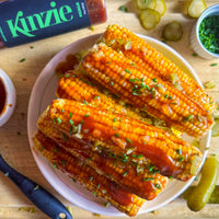 Corn on the cob with barbecue sauce and green onions on a white plate, with Kinzie pickles in the background.