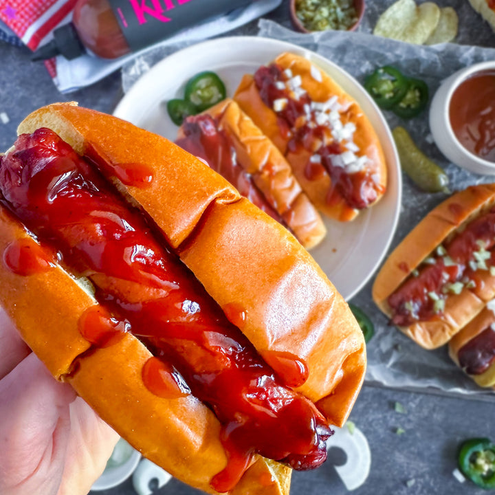 Hot dog with ketchup in a bun held by a hand, with more hot dogs and condiments in the background.