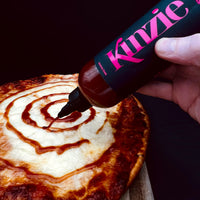 Hand using a bottle labeled 'Kinzie' to drizzle sauce on a pizza.