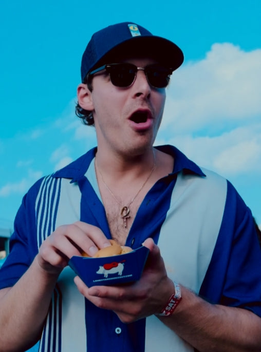 Man wearing sunglasses and a blue cap, holding a bowl with food against a blue sky.